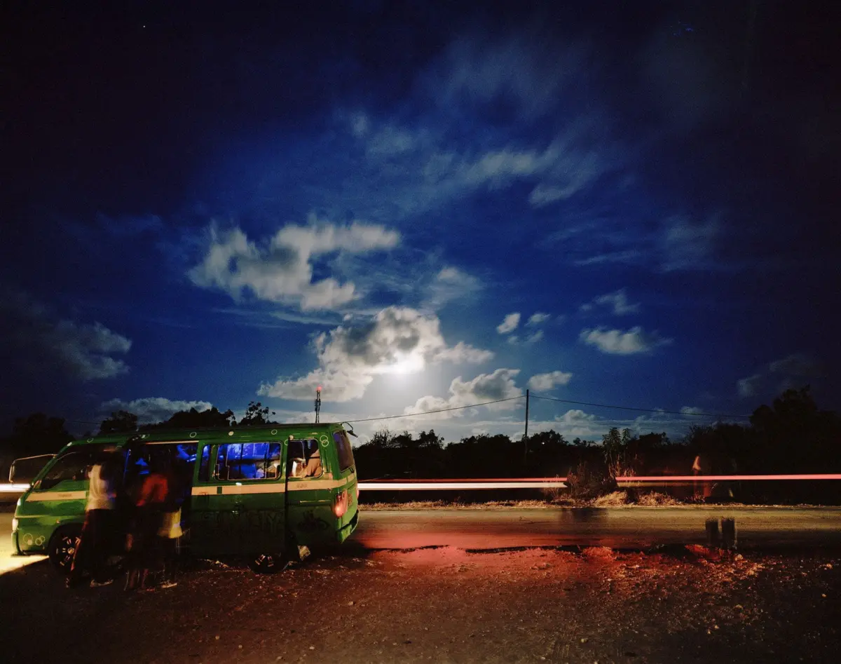 Green passenger van with open door at night; several people by roadside, blue interior light, moonlit sky, red light trail across road.