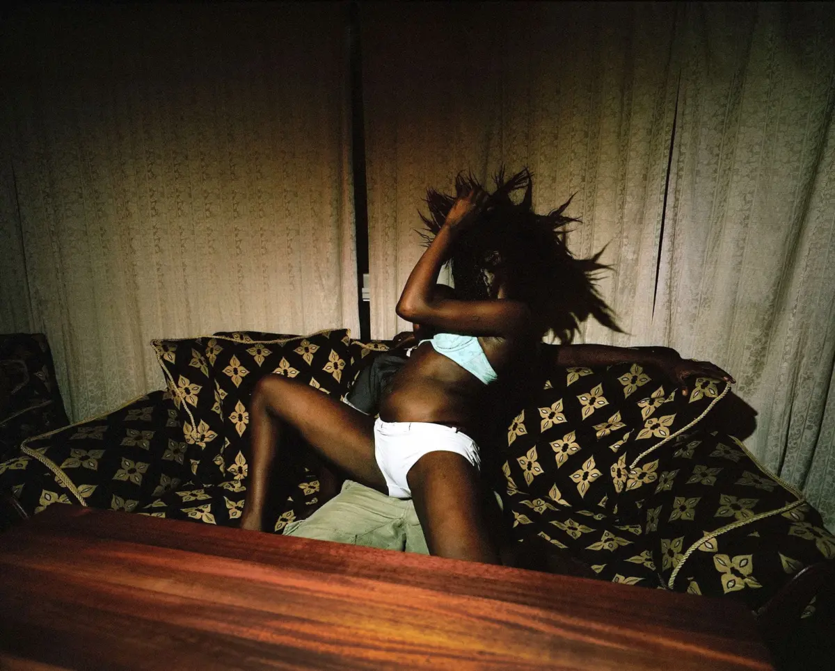 Person on a floral-patterned couch dancing, hair flying, wearing white underwear and light shorts in a dim living room.