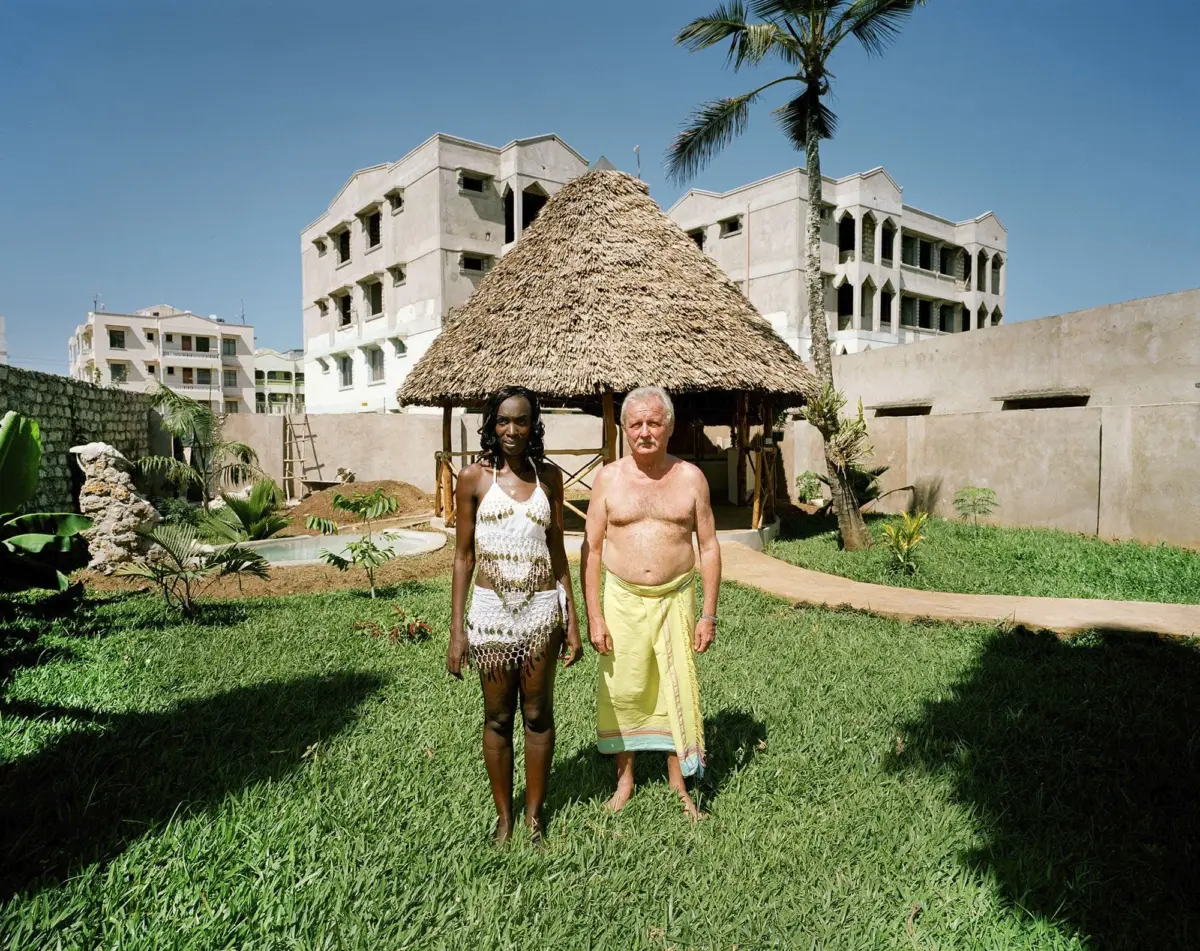 Two adults stand in a grassy yard near a thatched-roof gazebo, with an unfinished concrete building and palm tree against a blue sky.