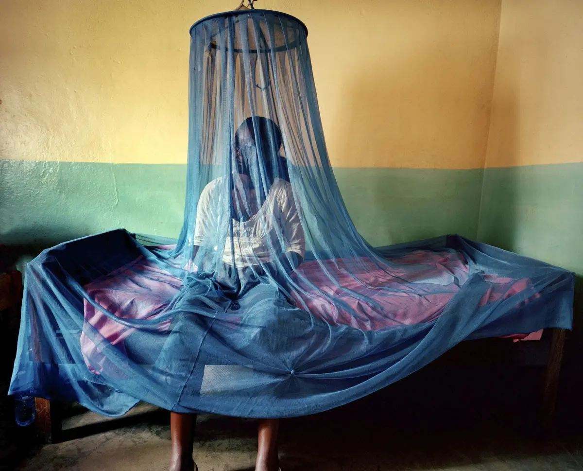 Person sits on bed beneath a draped blue mosquito net in a yellow-green room.