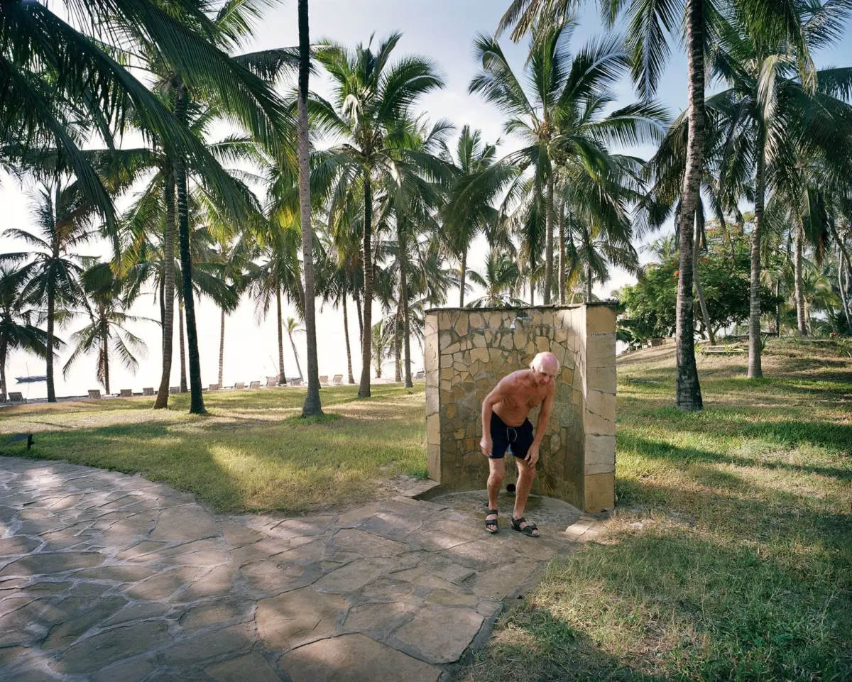 Shirtless adult person leaning from a stone pillar on a tropical beach amid palm trees.