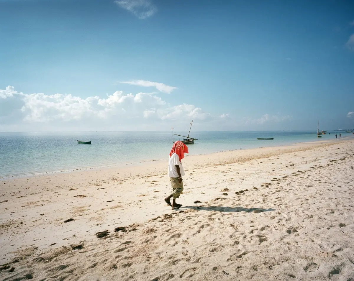 Person walking along sandy beach near calm blue sea; red cloth draped over head, small boats near shore.