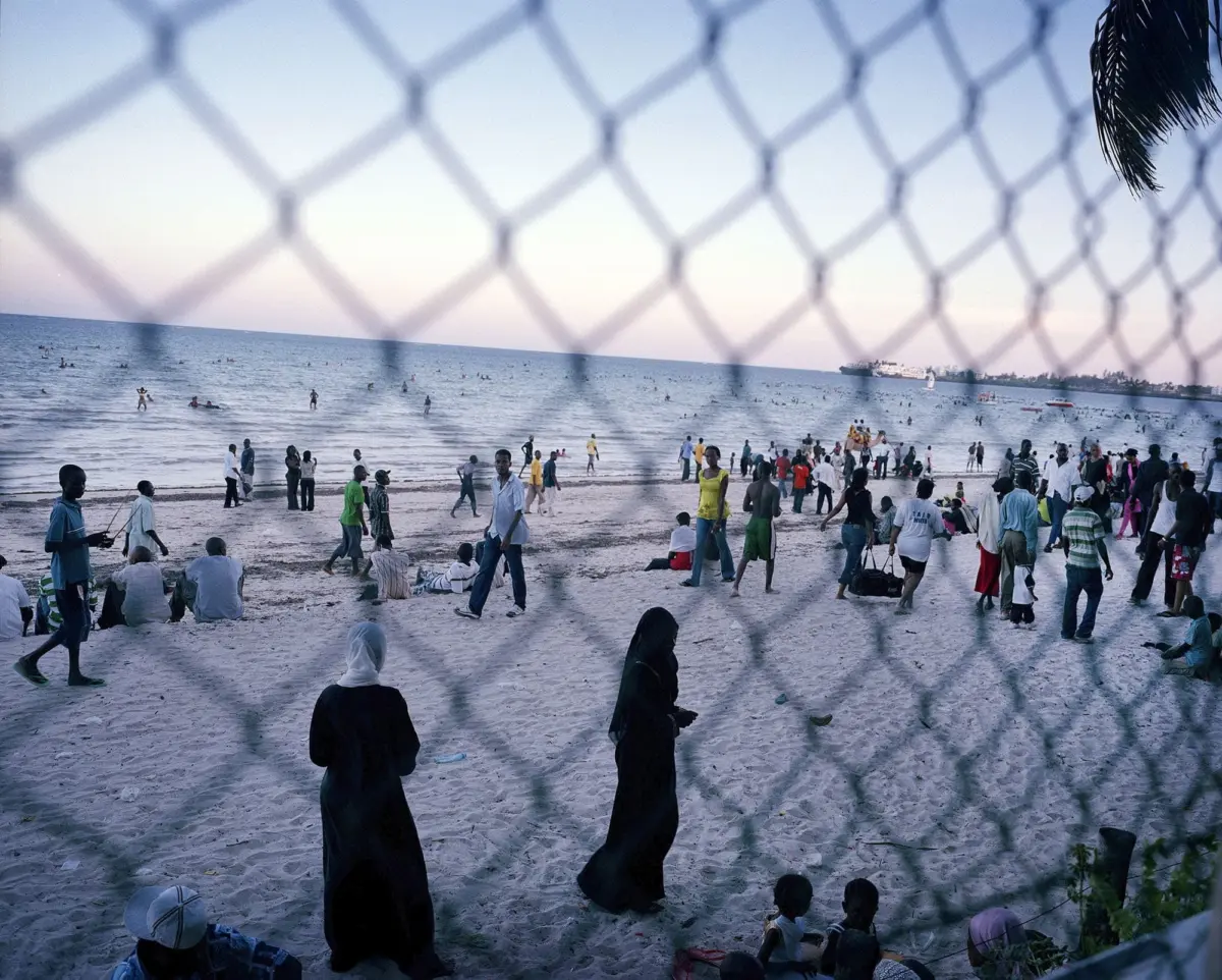 Crowded beach with people walking and swimming, viewed through a blurred chain-link fence toward the ocean.