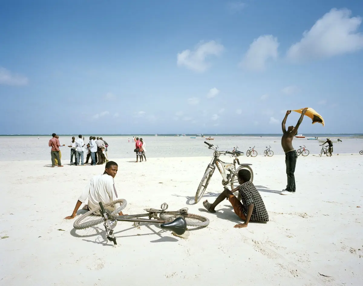 Two men and a boy sit near bicycles on a white-sand beach; a shirtless man raises an orange cloth while a distant group gathers by the ocean.