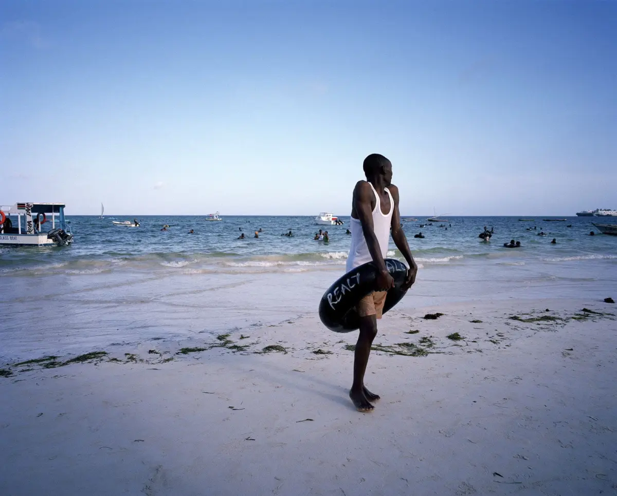 Person wearing a white tank top stands on a sandy beach, holding a black inflatable swim ring; sea with boats and swimmers in the background.