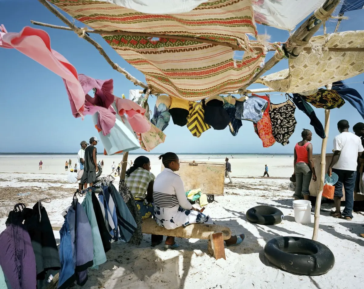 Beachside clothing stall with colorful garments hanging on a wooden clothesline under a fabric canopy; people sit and stand on white sand by the sea.