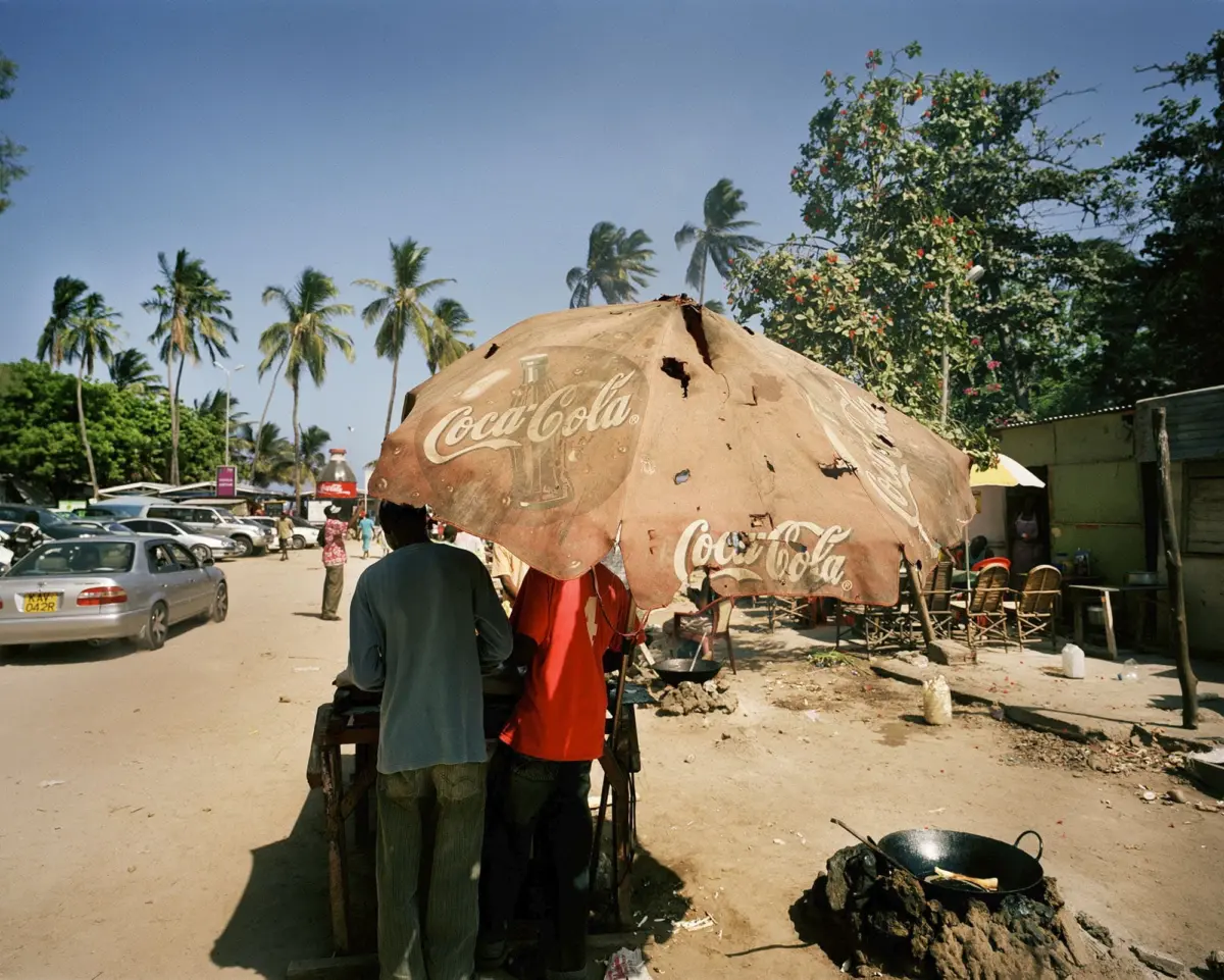 Tattered Coca-Cola umbrella shelters a street vendor stall as people gather in a tropical market with palm trees and parked cars.