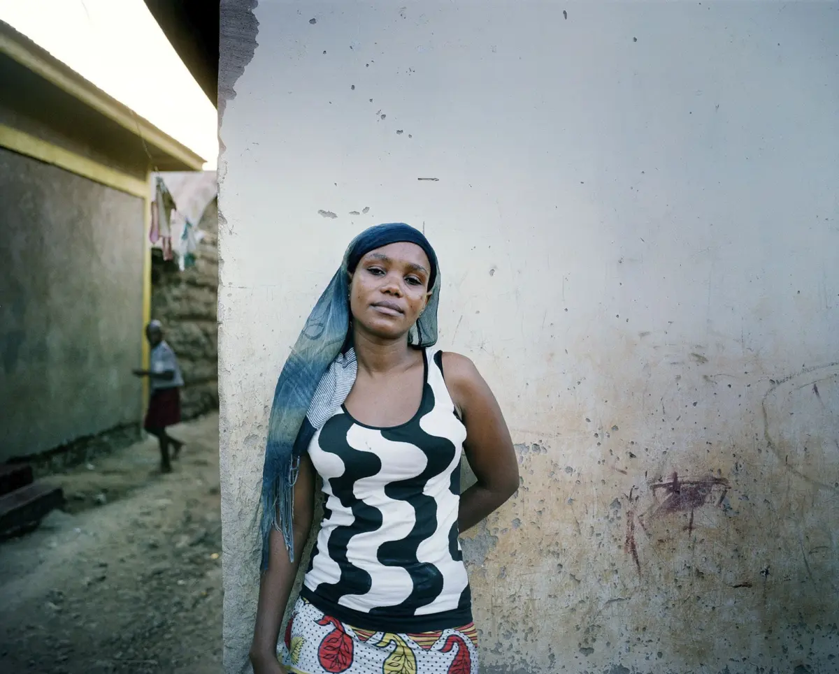 Person wearing blue headscarf and black-and-white zigzag dress standing against a weathered wall in an outdoor alley.