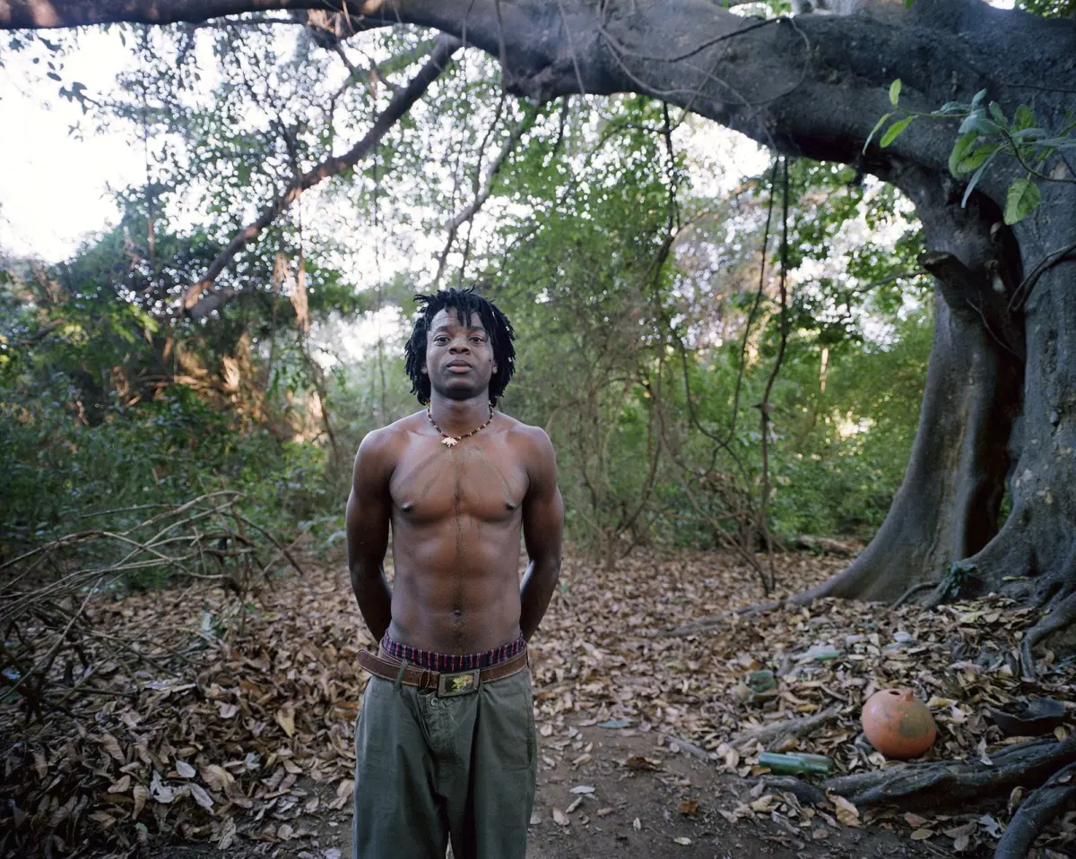 Shirtless young person with dreadlocks stands with hands behind back in a leaf-strewn forest clearing.