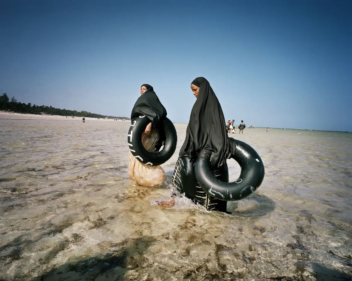 Two people wearing black hijabs wading in shallow beach water, each holding a black inflatable inner tube.