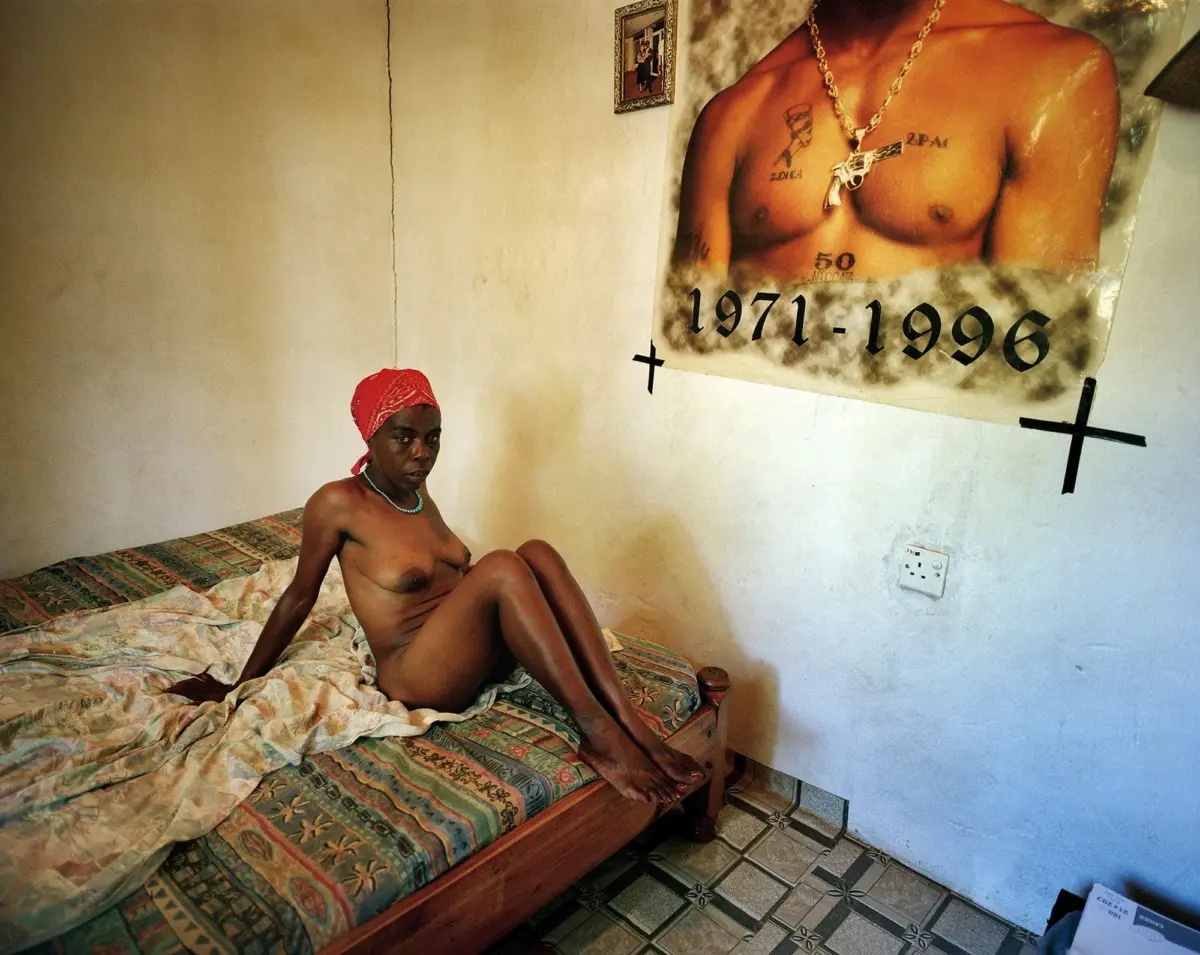 Nude person sits on a patterned bed in a bare room; red bandana and turquoise necklace; large poster of shirtless person with 1971-1996 on wall.