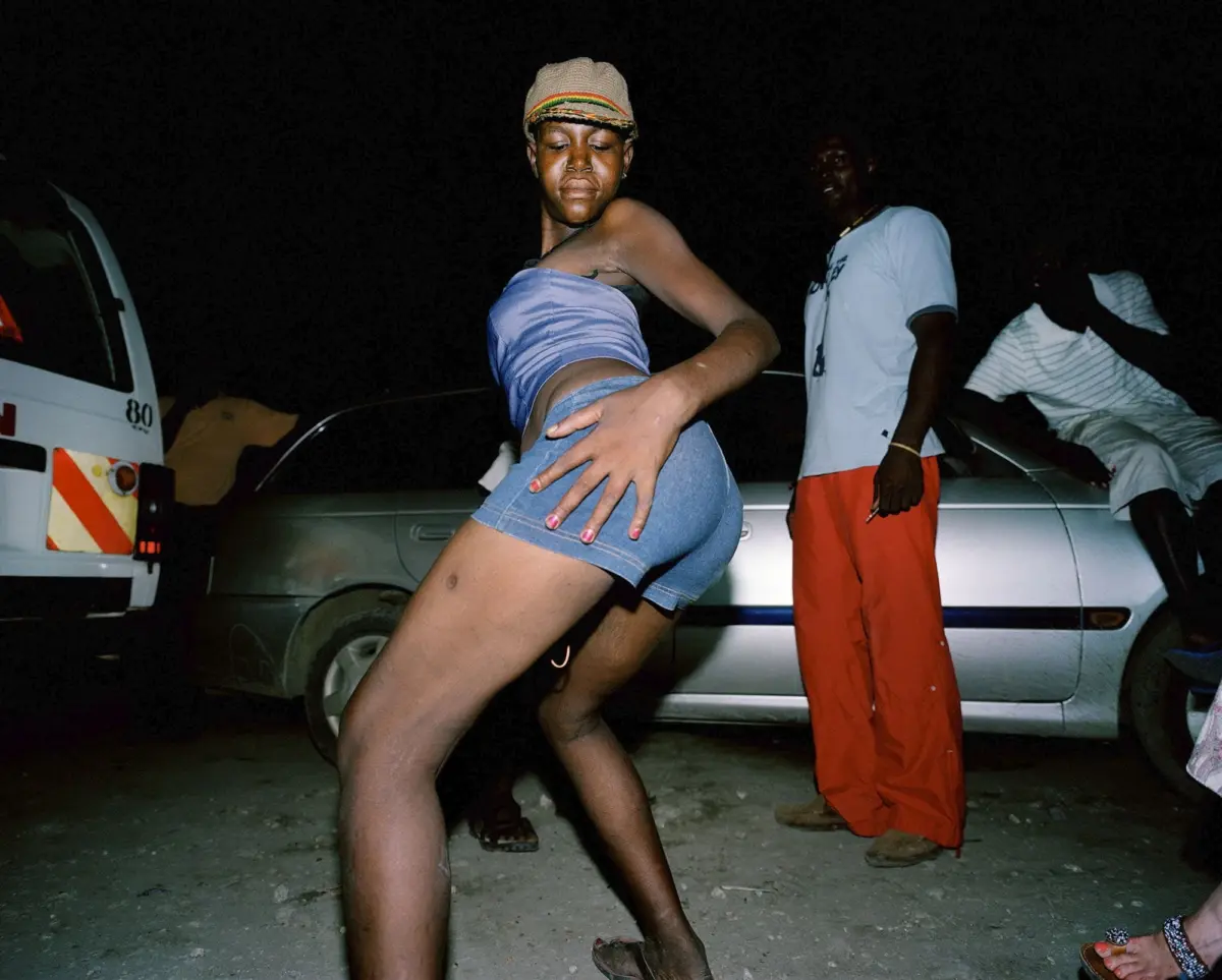 Person dancing in a blue tube top and denim shorts at night, in a dusty outdoor lot beside parked cars.
