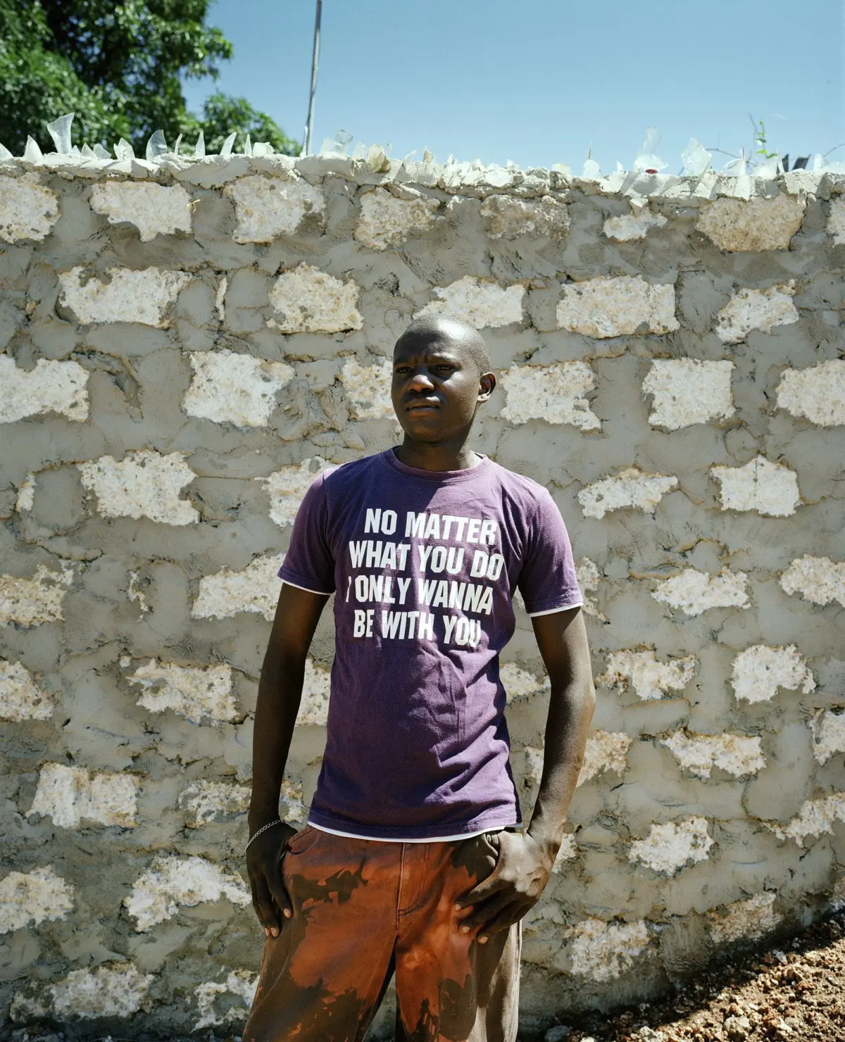 Adult person standing in front of a rough stone wall, wearing a purple T-shirt with white slogan and orange-brown shorts.