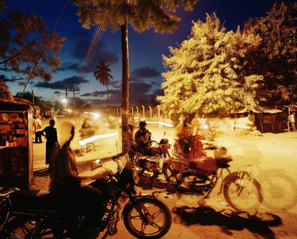 Night street scene with many motorcycles and pedestrians, orange glow from streetlights, tropical trees lining the road.