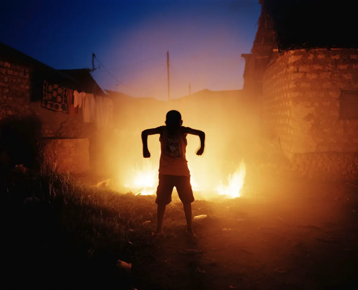 Silhouette of a child with bent arms standing before two fires on a dirt village street at dusk, flanked by stone houses.