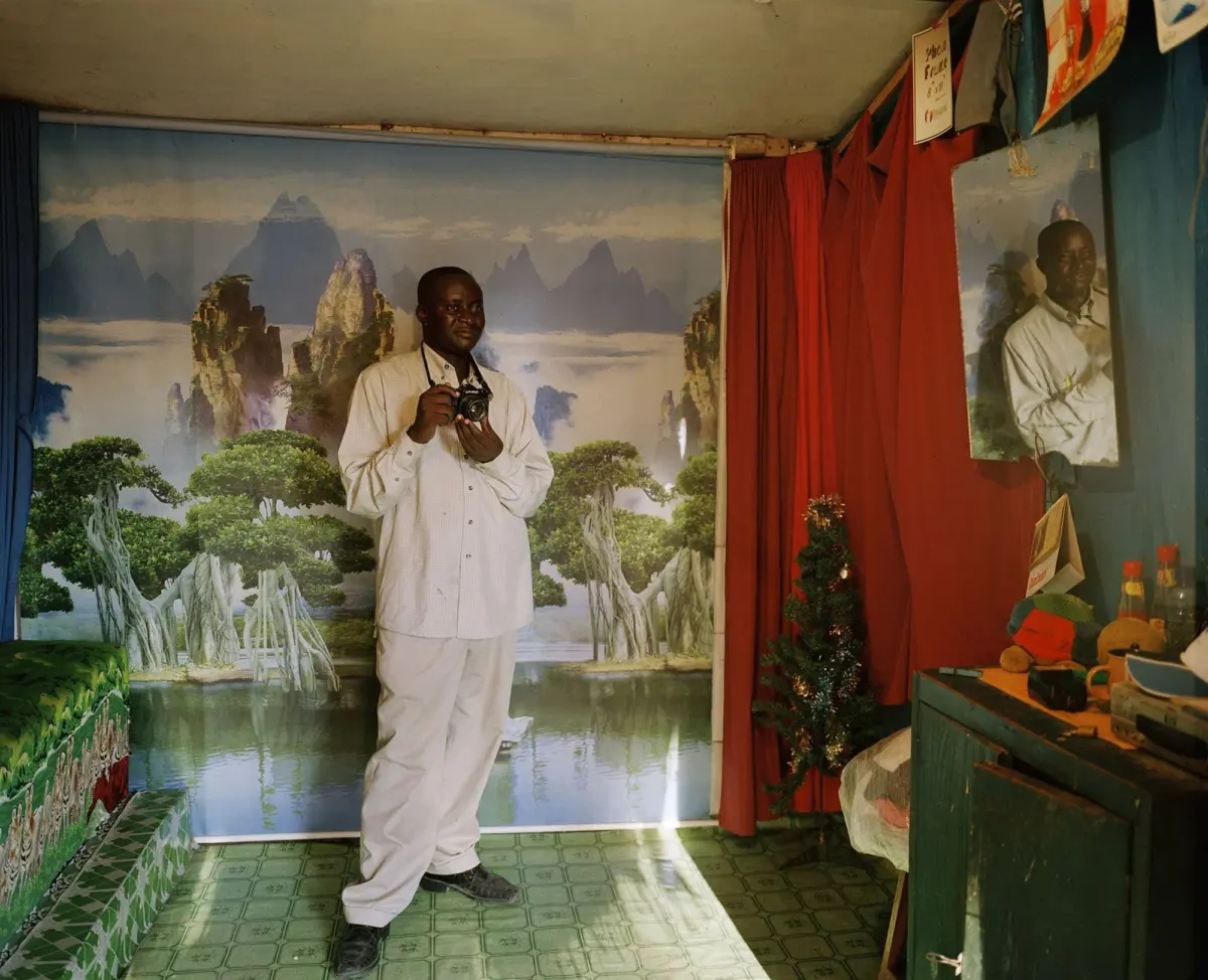 Person in light shirt holds a camera, standing in front of a landscape mural with mountains and trees, red curtains on the right.