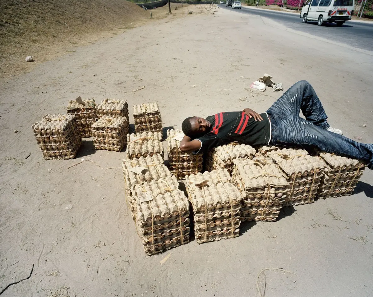 Man lying on stacked egg cartons along a dusty roadside, with vehicles visible in the background.