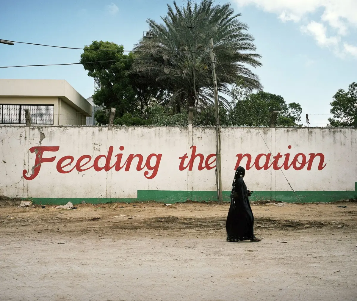 Person in black abaya walks past a white wall with red cursive 'Feeding the nation' under a blue sky; palm trees and power lines in background.