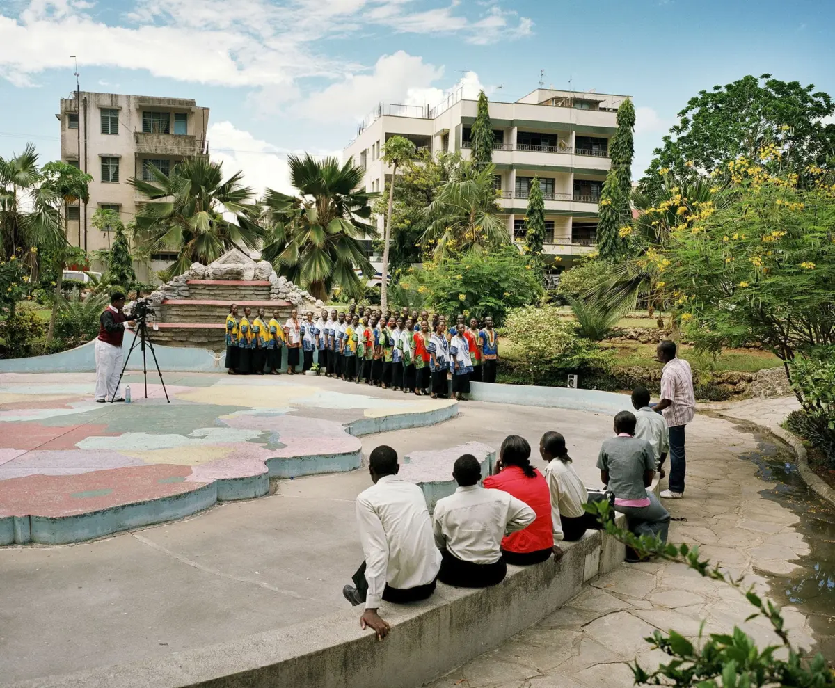 Choir of students in colorful vests sings in an outdoor courtyard while a cameraman films, with spectators seated on a curved wall.