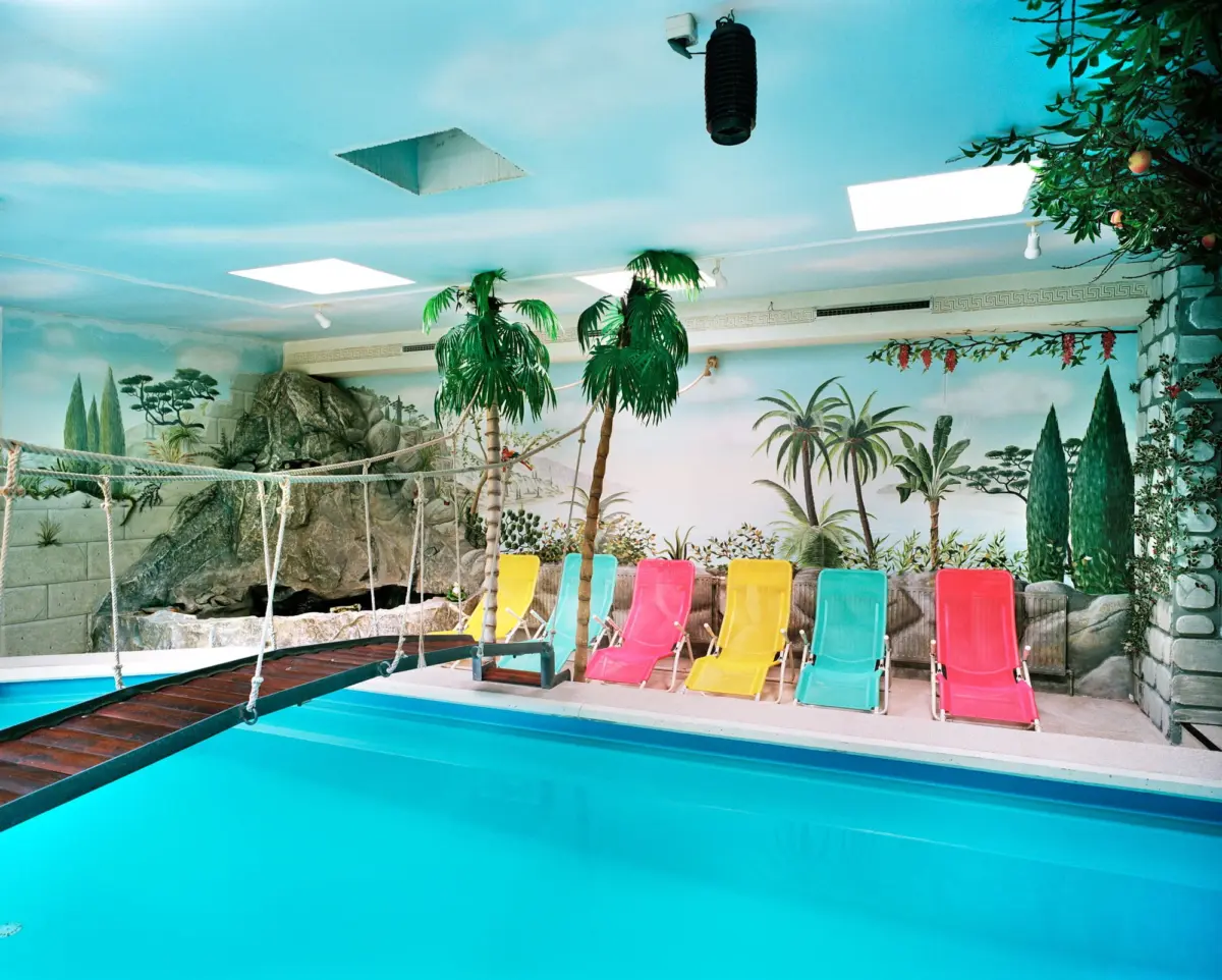 Indoor tropical pool area with turquoise water, vibrant plastic lounge chairs, palm trees, and a mural of a jungle landscape.