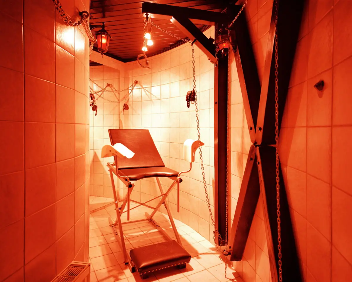 Red-lit dungeon room with a padded bondage chair, hanging chains, pulleys, and wooden supports along tiled walls.