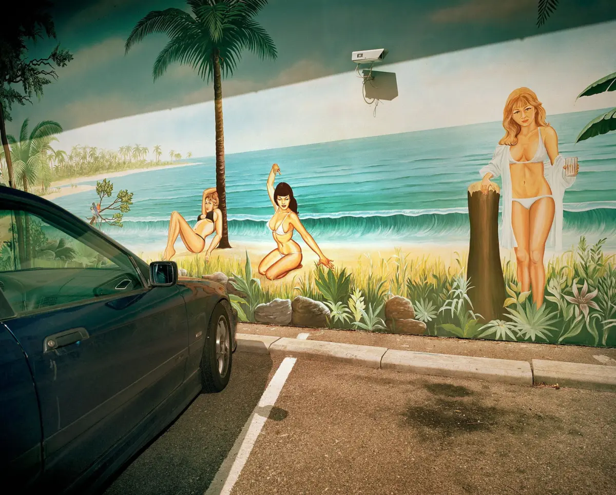 Dark green sedan parked in a parking lot in front of a tropical beach mural with bikini-clad women and palm trees.