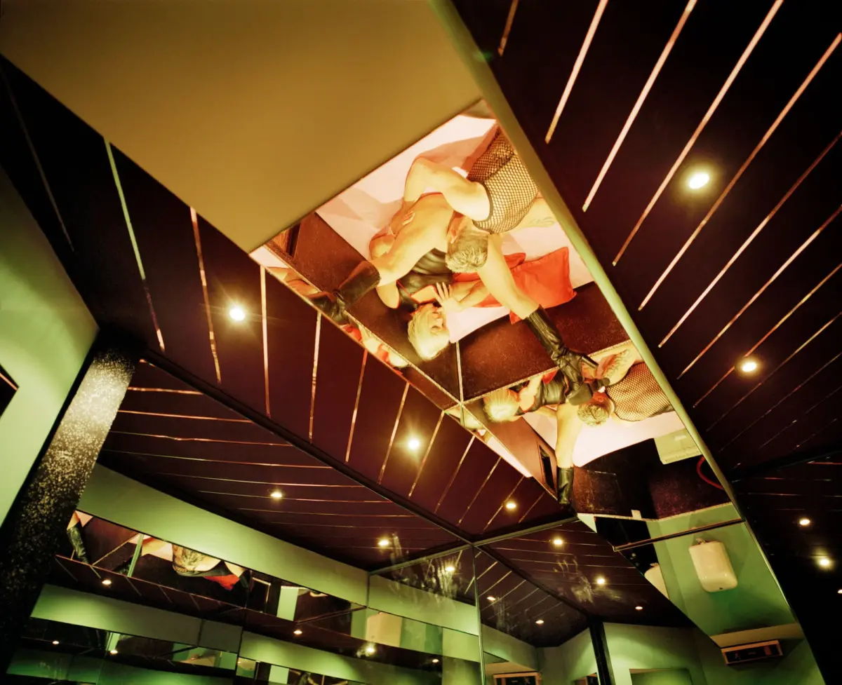 Two performers in a glass display booth on a padded bed, wearing fishnet stockings, inside a green-lit nightclub with angled panels.