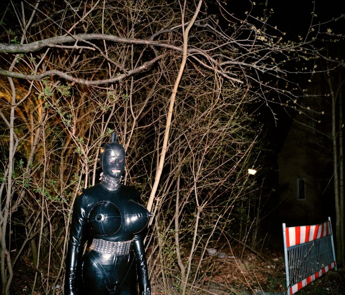 Person in a black Batman costume stands outdoors at night among bare tree branches; construction barrier visible to the right.