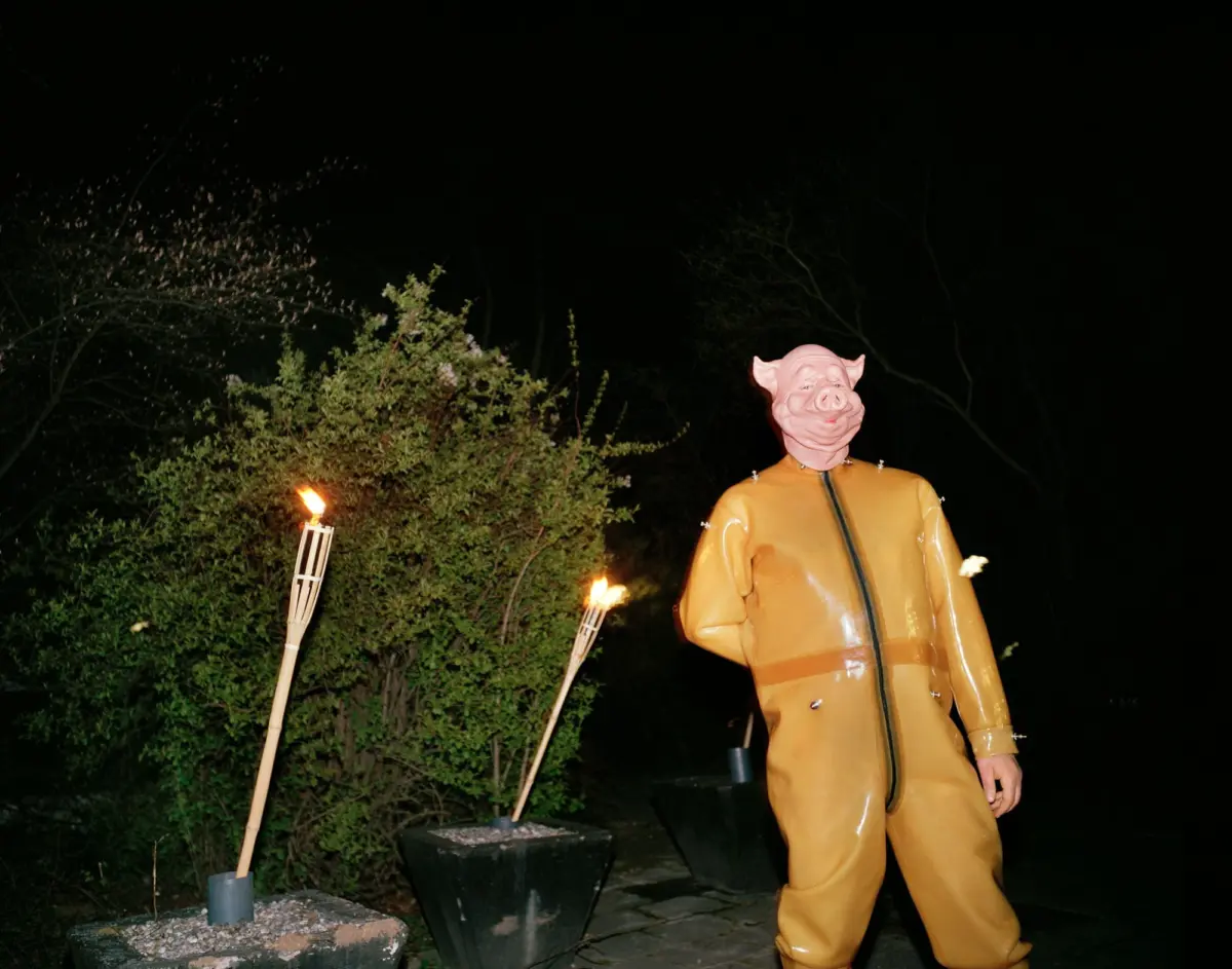 Person wearing a pig mask in a yellow hazmat suit outdoors at night, beside tiki torches and shrubs.