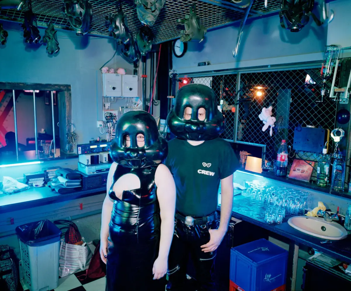 Two people in shiny black latex outfits wear large gas-mask helmets in a high-tech bar-lab with hanging masks and glassware.