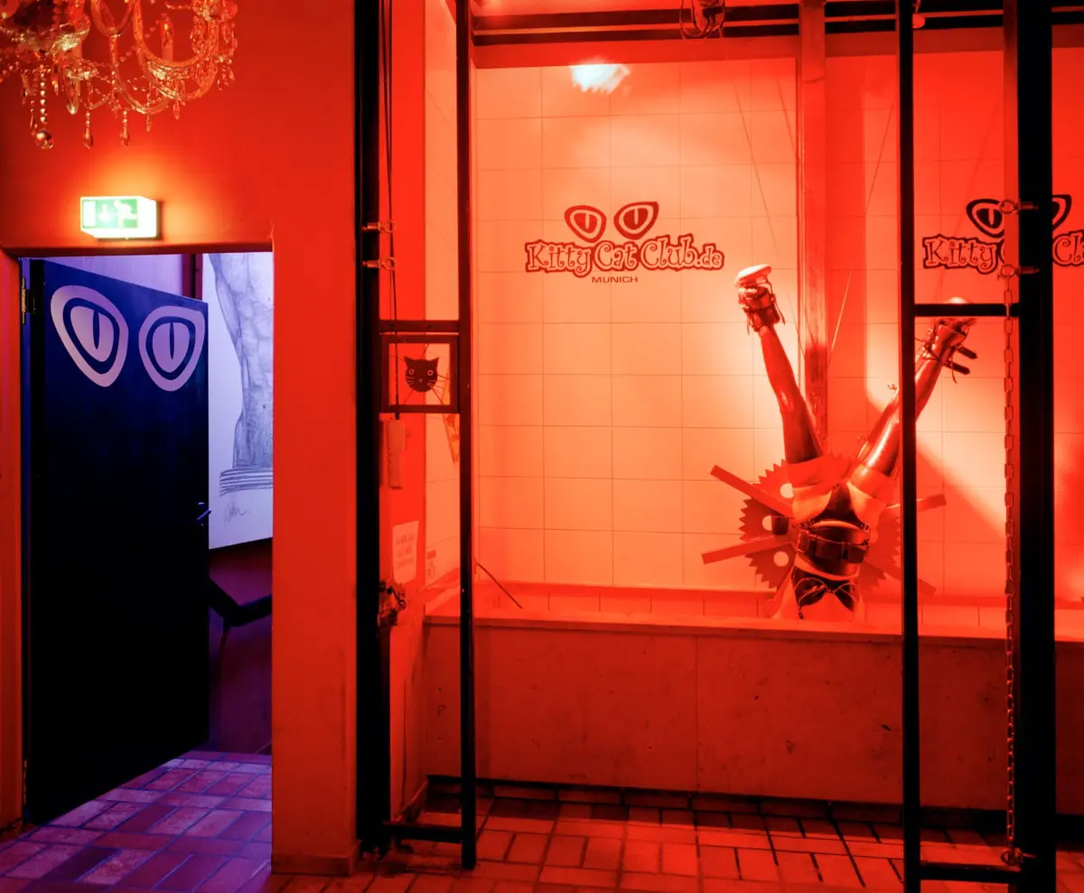 Red-lit storefront display window showing a mannequin leg and the KittyCatClub.de Munich logo on tiled wall.