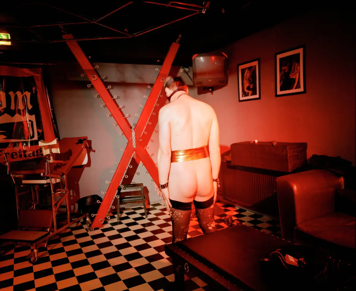 Shirtless person in metallic belt and stockings stands with back to camera in a red-lit BDSM room; bondage frame, checkered floor.