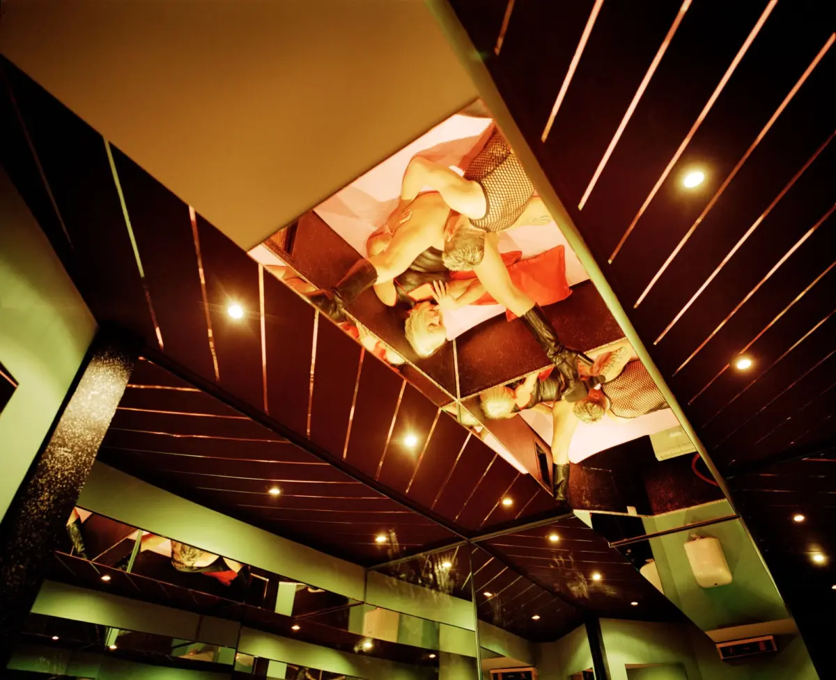Group of performers on a pink, cushion-covered stage inside a geometric nightclub; one wears fishnet shorts and boots.