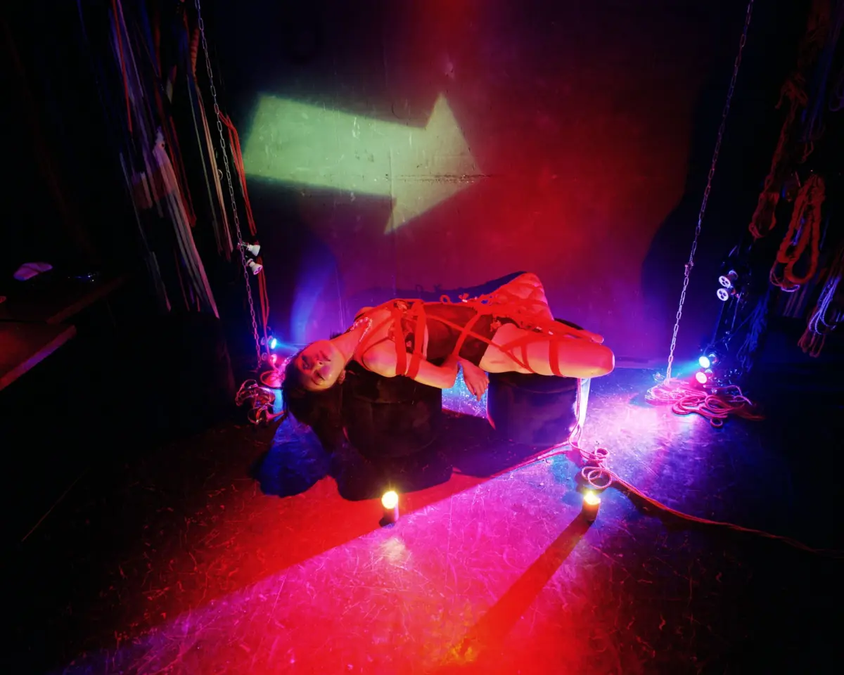 Person tied with red ropes on a dark stage amid hanging ropes and chains, lit by colorful stage lighting.