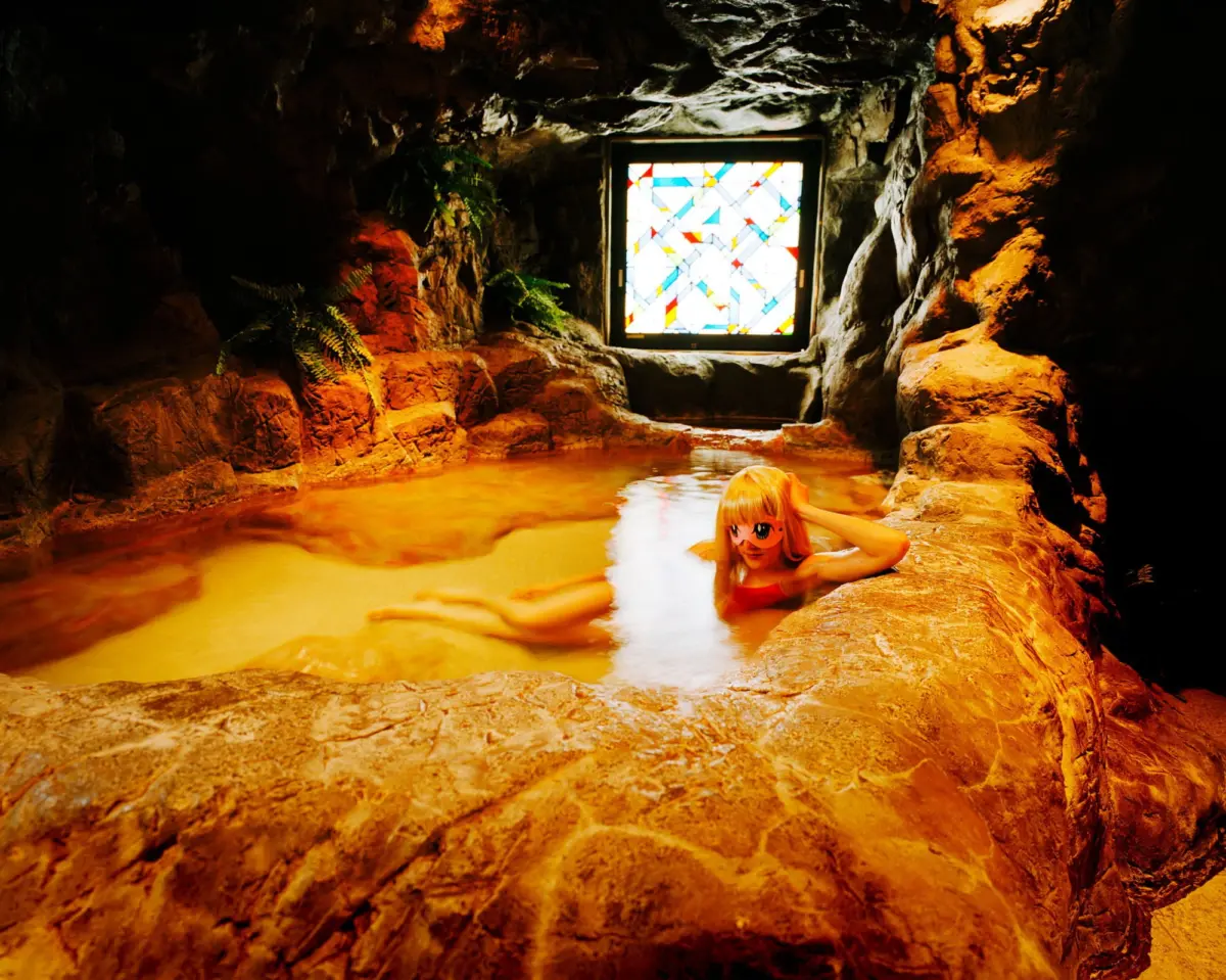 Person with orange hair wearing a mask floats in a rocky underground pool, with a colorful stained-glass window in the cave wall.