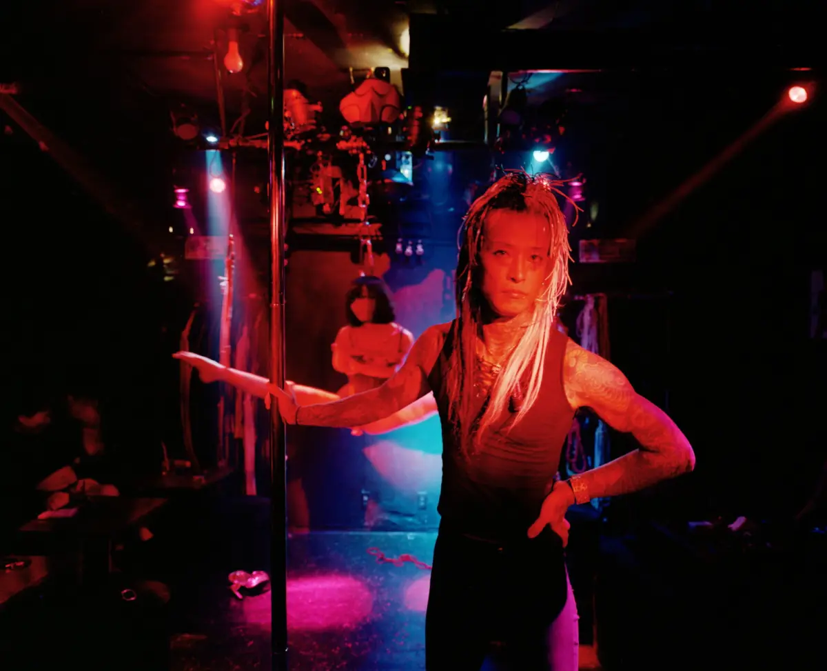 Pole dancer with braided hair and tattoos performing on a dim club stage under red and blue lights.