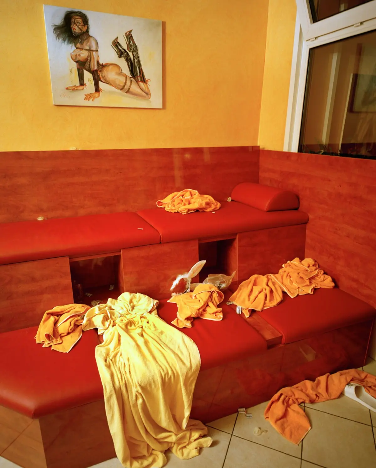 Red-orange sectional sofa with scattered towels in a warm room; painting of a bound nude figure on the wall near a window.