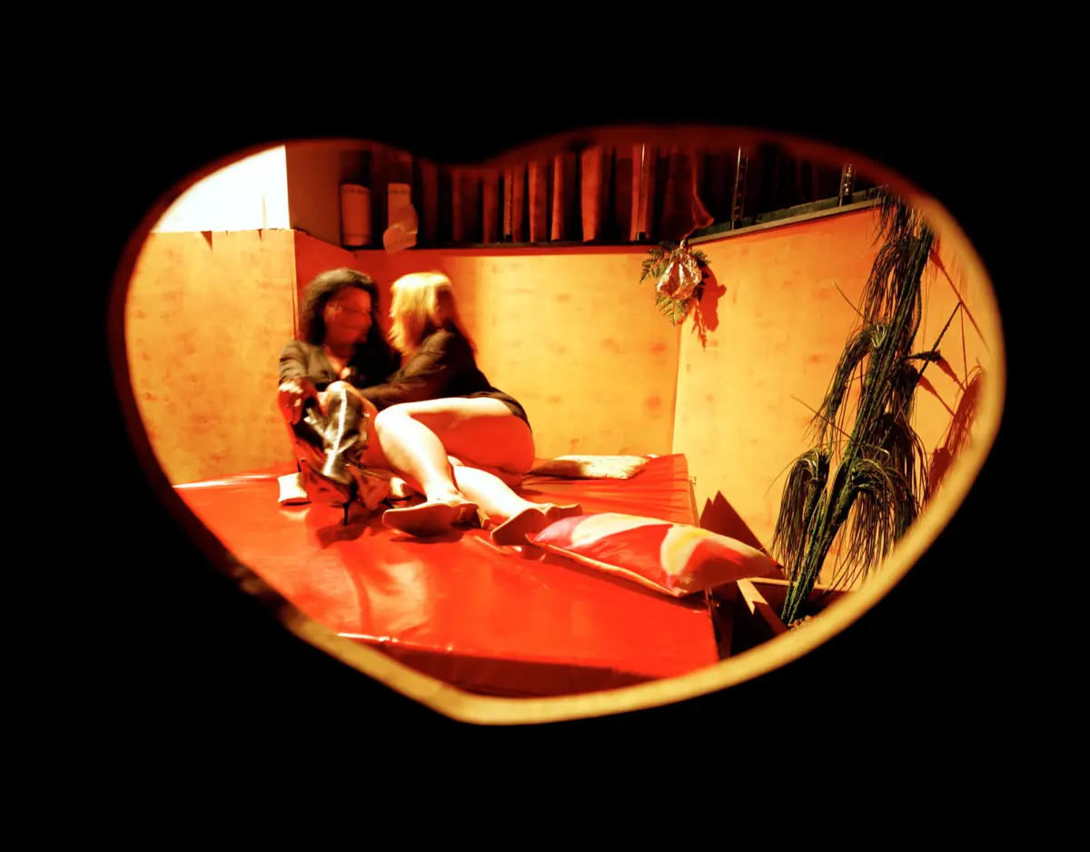 Two people embracing on a red sofa, in a warmly lit orange room, viewed through a circular window.