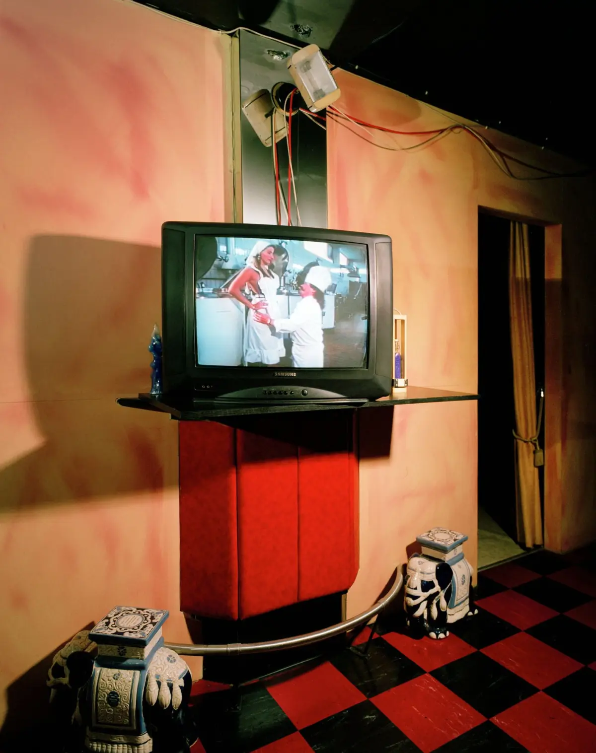 Vintage Samsung CRT TV on a red cabinet shows two people in a kitchen; pink walls, exposed wiring, and ceramic foo dogs nearby.