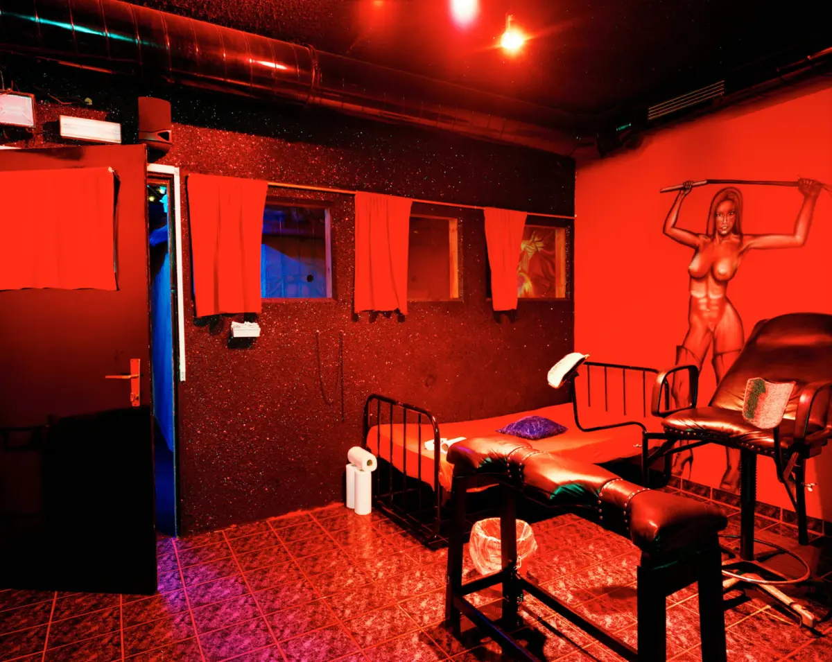 Red-lit BDSM dungeon with glittery walls, metal beds, red curtains, and a mural of a nude figure raising a rod.