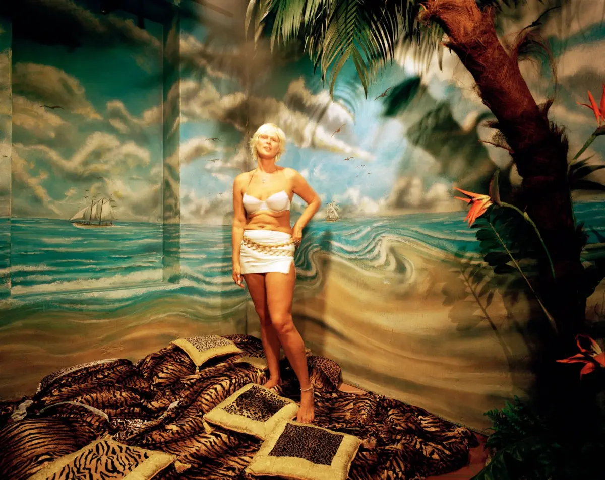 Blonde woman in white bikini top and mini skirt with chain belt poses in tropical mural scene with palm trees and sea, tiger-pattern cushions.