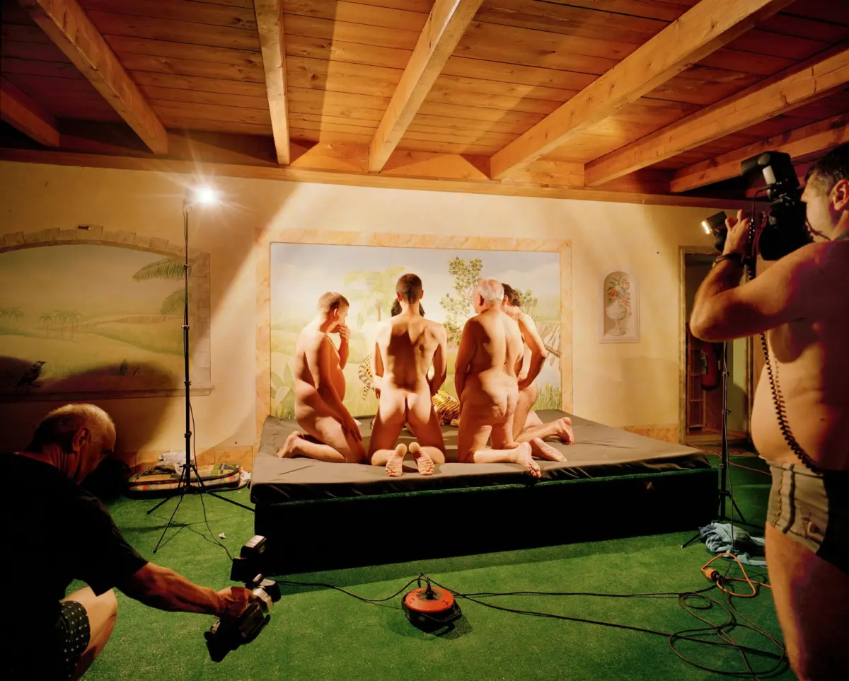 Nude adult men posing around a tropical mural on a raised platform while a photographer shoots them in a wood-beamed studio.