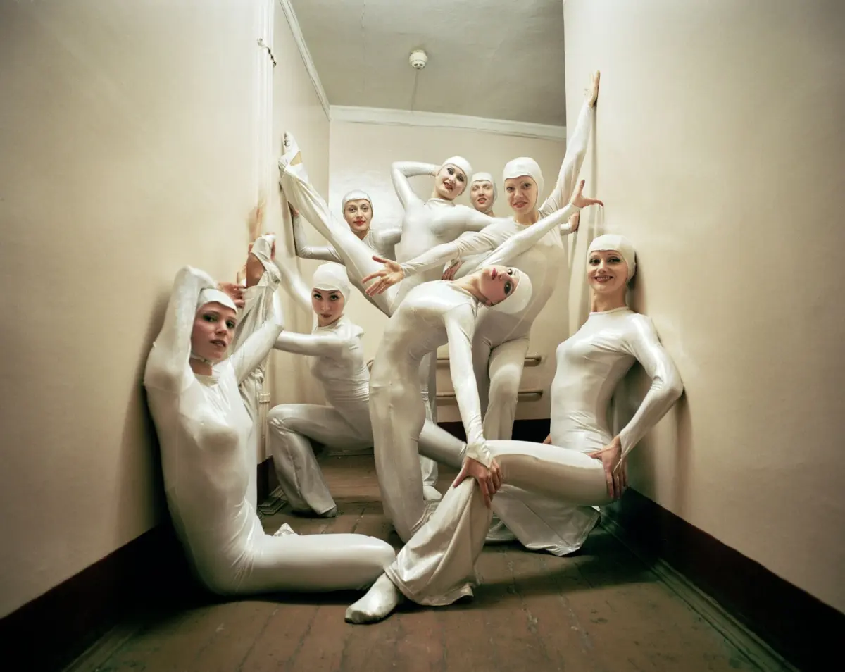 Group of dancers in white unitards in a narrow hallway, posing with extended limbs.