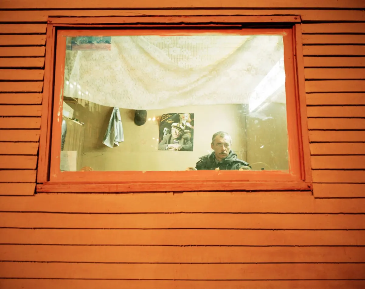 Adult with mustache seated at a desk inside a room, seen through an orange-framed window; posters on the wall and a hanging shirt.