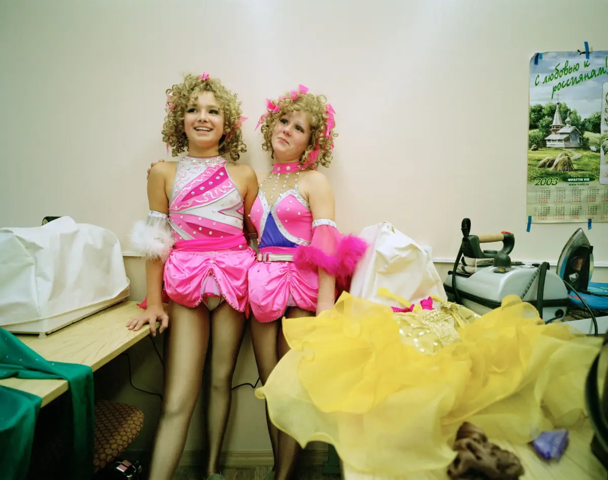 Two young girls with curly blonde hair in pink sequined dance costumes pose together in a backstage dressing room.