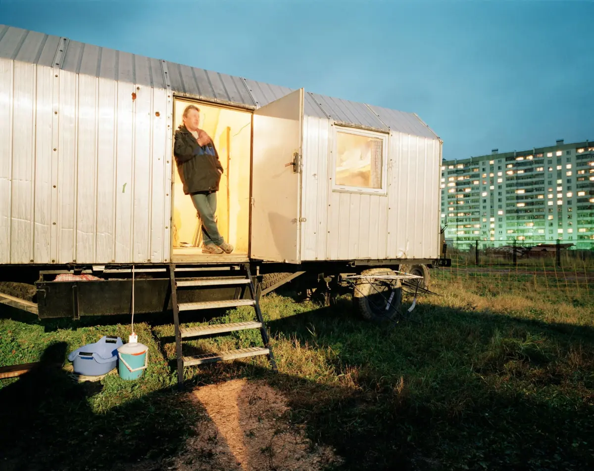 Person standing in the doorway of a metal mobile trailer on wheels in a grassy field, with a residential complex in the background.