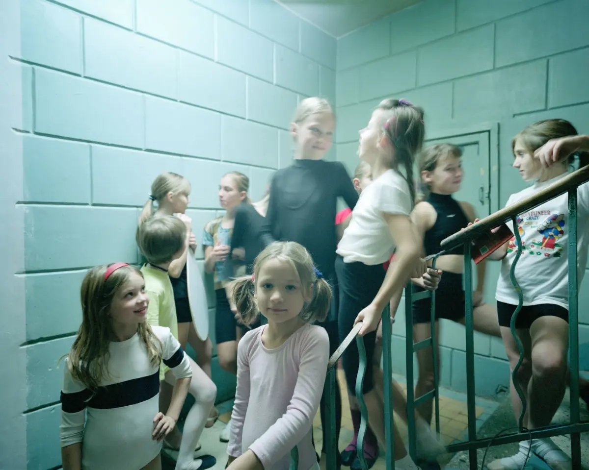 Group of children in leotards in a teal-walled stairwell; central child faces the camera.