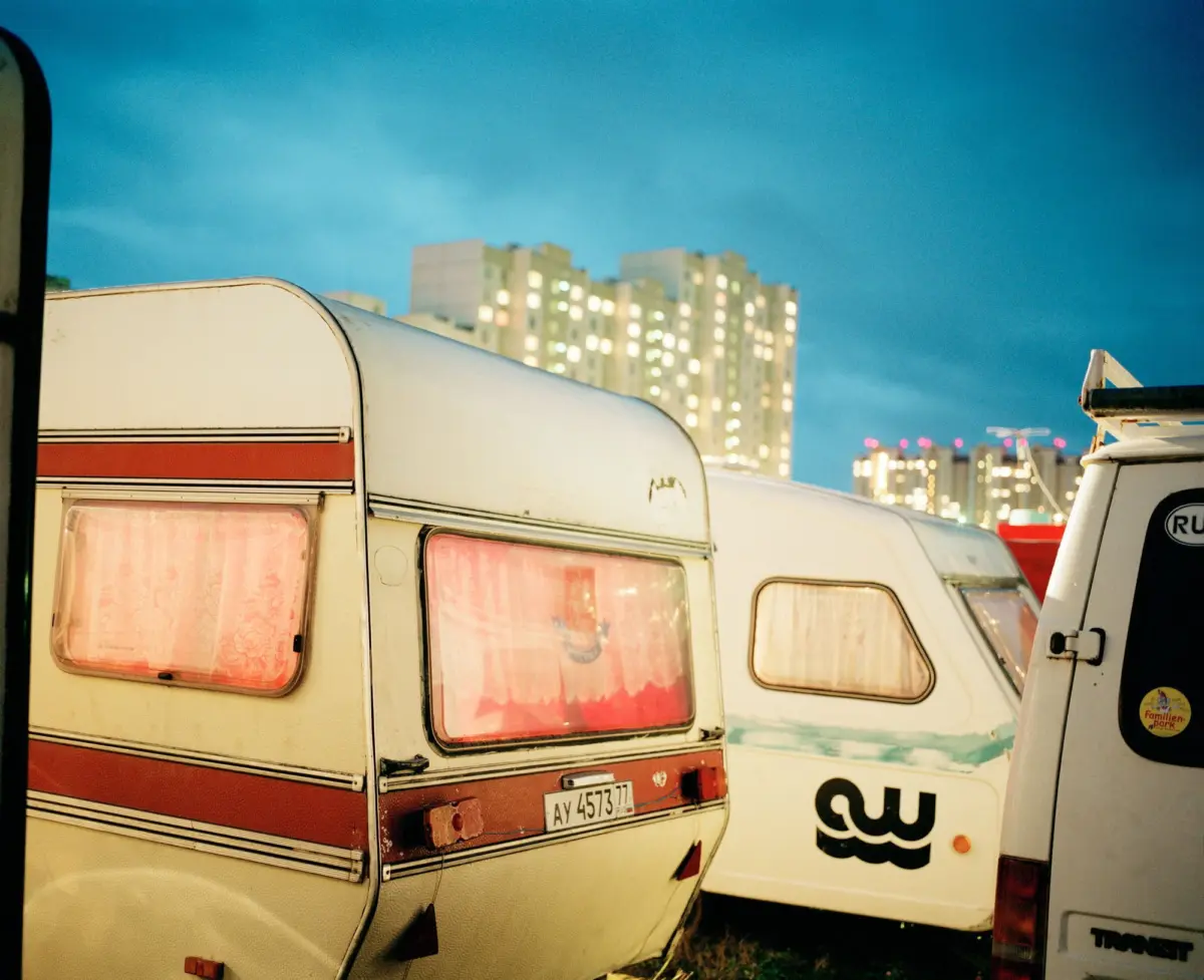 Row of vintage caravans parked in a lot, pink-curtained windows, with city skyline at dusk.