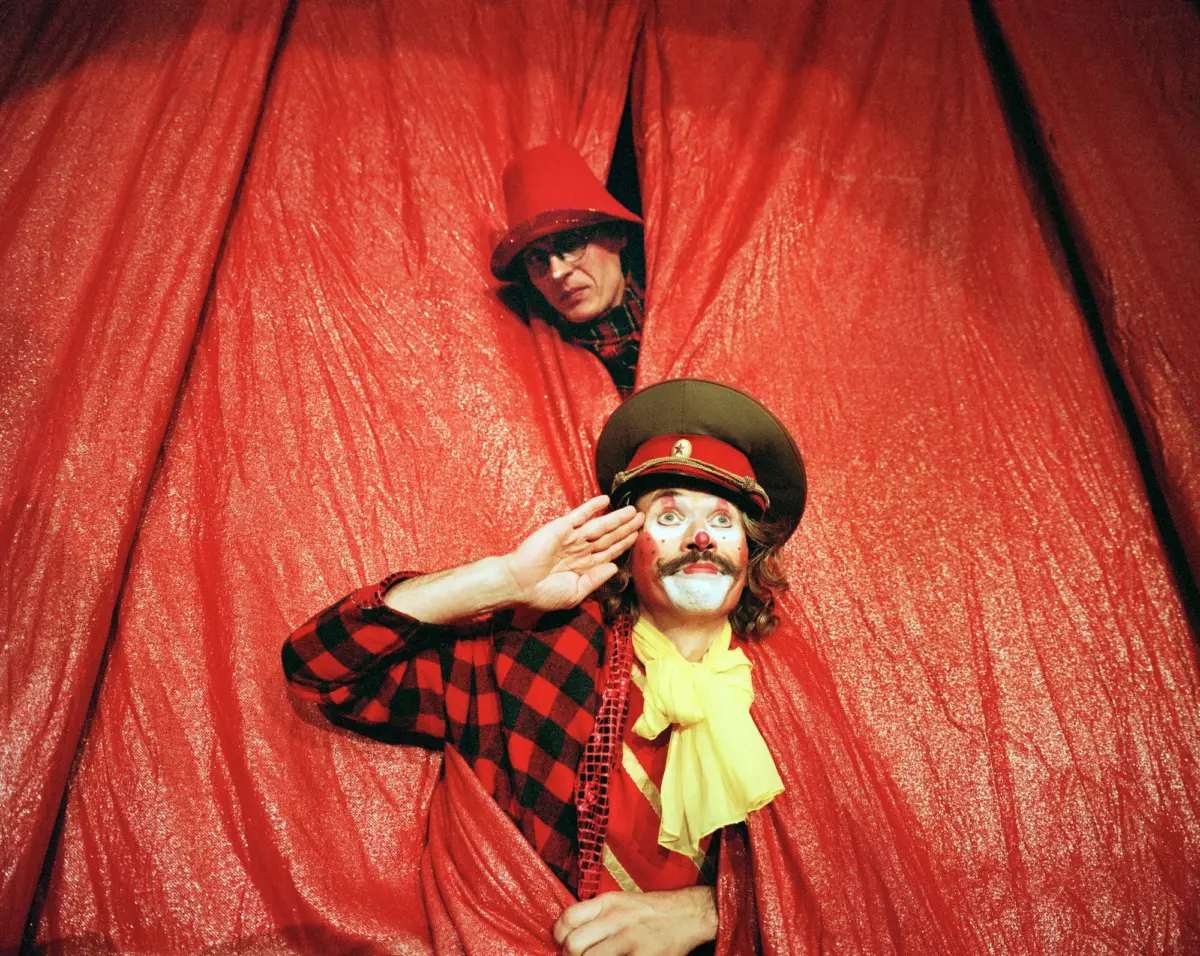 Two clowns peek from behind a red velvet curtain; one in a red hat and sunglasses, the other in a green hat with painted face and yellow scarf.