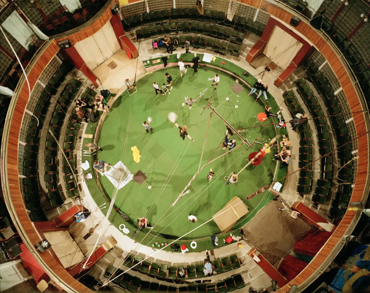 Top-down view of a circular circus arena with a green floor, performers practicing amid rigging and crew, surrounded by tiered seating.